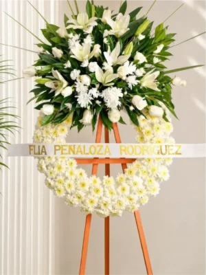 Corona Fúnebre Espinas de Consuelo con base de margaritas blancas y copete de lirios, rosas y follaje, sobre atril de madera.