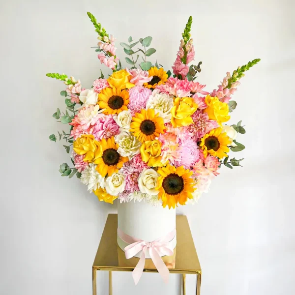 Ramo de girasoles con rosas blancas y amarillas, flores rosadas y follaje en base cilíndrica con lazo rosado.