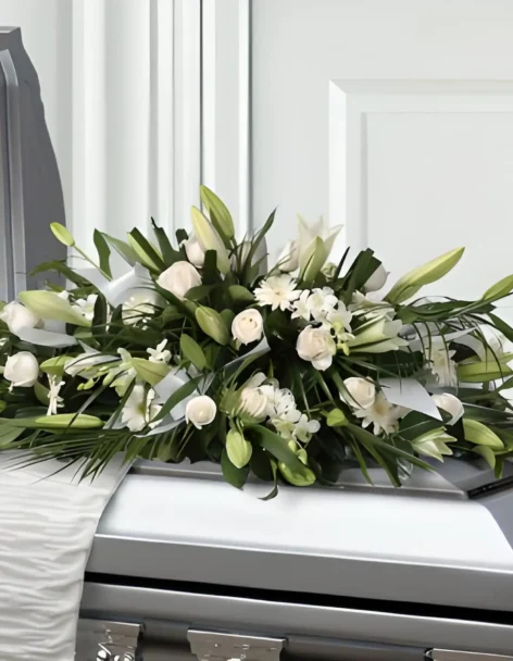 Cubre cofre de rosas y lirios blancos con follaje verde, extendido sobre un ataúd plateado.