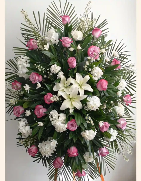 Pedestal fúnebre en forma de abanico con hojas de palma, rosas fucsia, hortensia y lirios blancos, cinta naranja.