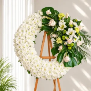 Corona Fúnebre Ágata Corona Fúnebre Ágata circular de crisantemos blancos con rosas crema, lirios blancos y follaje verde, sobre atril de madera.