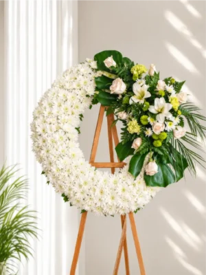 Corona Fúnebre Ágata circular de crisantemos blancos con rosas crema, lirios blancos y follaje verde, sobre atril de madera.