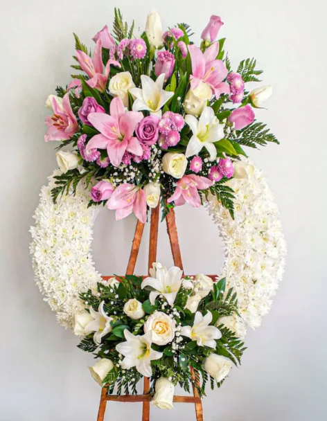 Corona fúnebre de dos niveles con lirios y rosas rosadas, follaje verde y base circular de crisantemos blancos