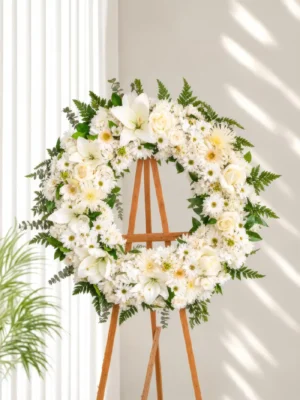 Corona Fúnebre Esmeralda de crisantemos, lirios y rosas blancas con gerberas araña y follaje verde, sobre atril de madera.