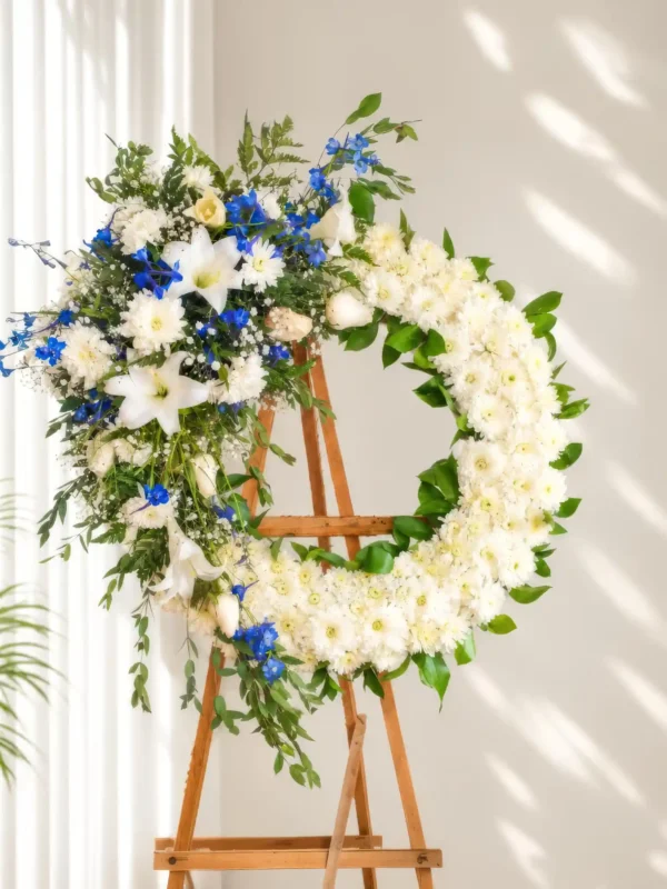 Corona Fúnebre Magia Eterna circular de crisantemos blancos con lirios y flores azules a un costado, sobre atril de madera.