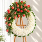Corona Fúnebre Memoria circular de crisantemos blancos con rosas color coral en la parte superior, sobre atril de madera.