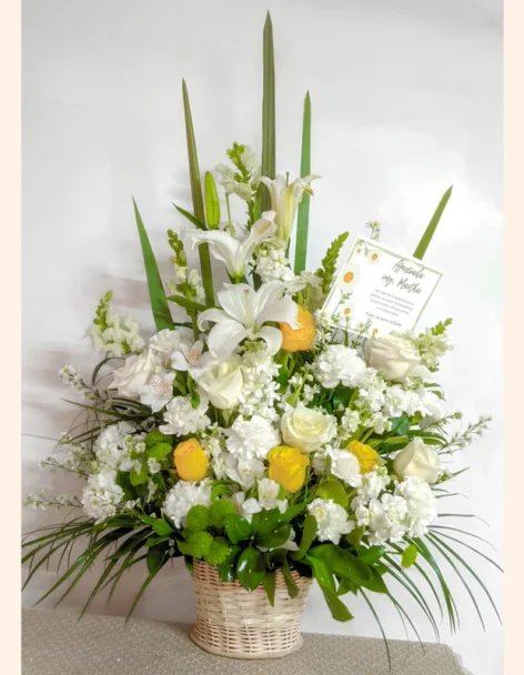 Arreglo fúnebre de lirios y rosas blancas y amarillas con tarjeta de condolencia, en cesta de mimbre.